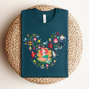 K&ouml;nnte beinhalten: T-Shirt in Teal mit einer farbenfrohen Grafik einer Meerjungfrau, eines Prinzen und anderer Charaktere, die in Form eines Mickey Mouse-Kopfes angeordnet sind. Das Design umfasst verschiedene Meereskreaturen und florale Elemente. Das Shirt wird auf einer gewebten Oberfl&auml;che pr&auml;sentiert.