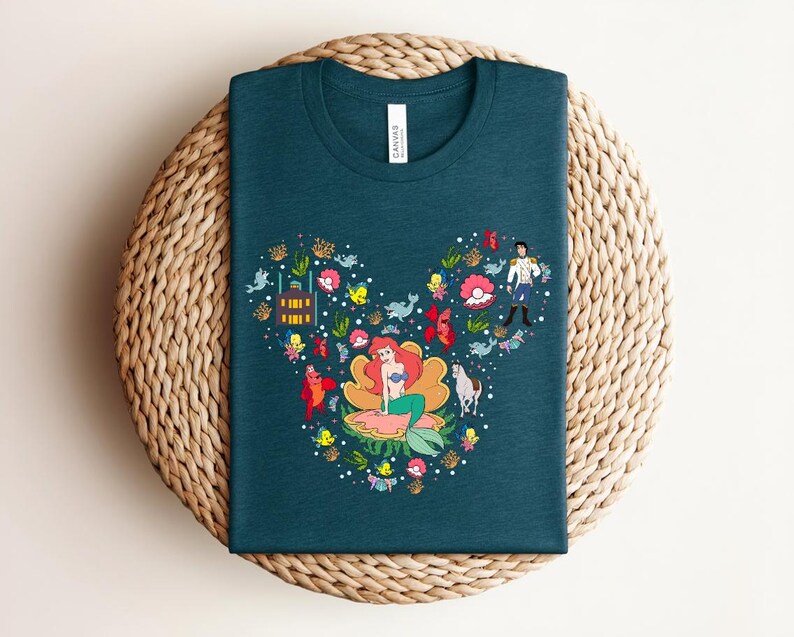 K&ouml;nnte beinhalten: T-Shirt in Teal mit einer farbenfrohen Grafik einer Meerjungfrau, eines Prinzen und anderer Charaktere, die in Form eines Mickey Mouse-Kopfes angeordnet sind. Das Design umfasst verschiedene Meereskreaturen und florale Elemente. Das Shirt wird auf einer gewebten Oberfl&auml;che pr&auml;sentiert.