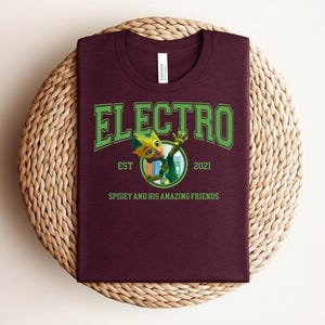 May include: A maroon t-shirt featuring the word "ELECTRO" in green, arched lettering. Below, a cartoon character is depicted within a circle, and the text "EST 2021" and "SPIDEY AND HIS AMAZING FRIENDS" are also present. The shirt is displayed on a woven, circular mat.
