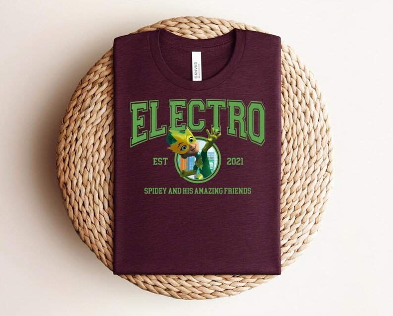May include: A maroon t-shirt featuring the word "ELECTRO" in green, arched lettering. Below, a cartoon character is depicted within a circle, and the text "EST 2021" and "SPIDEY AND HIS AMAZING FRIENDS" are also present. The shirt is displayed on a woven, circular mat.