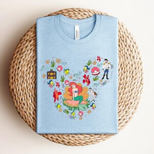 K&ouml;nnte beinhalten: Hellblaues T-Shirt mit einer farbenfrohen Grafik von Arielle aus Arielle, die Meerjungfrau, in Form eines Mickey Mouse-Ohrs angeordnet. Das Design umfasst florale Elemente, Geb&auml;ude und Charaktere aus dem Film. Das Shirt besteht aus weichem Material und hat einen Rundhalsausschnitt.