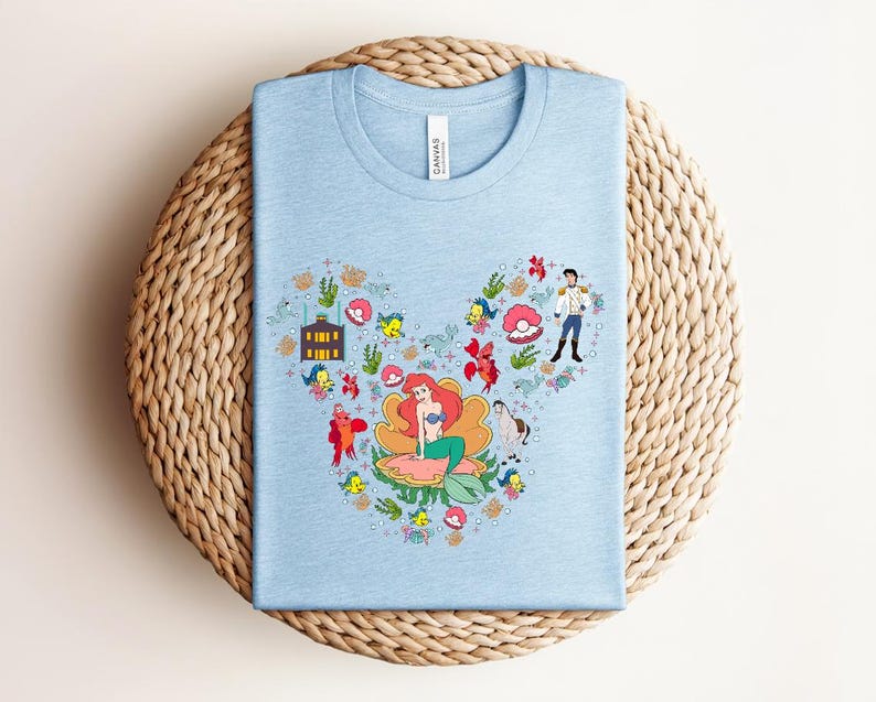 K&ouml;nnte beinhalten: Hellblaues T-Shirt mit einer farbenfrohen Grafik von Arielle aus Arielle, die Meerjungfrau, in Form eines Mickey Mouse-Ohrs angeordnet. Das Design umfasst florale Elemente, Geb&auml;ude und Charaktere aus dem Film. Das Shirt besteht aus weichem Material und hat einen Rundhalsausschnitt.