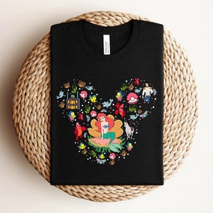 K&ouml;nnte beinhalten: Schwarzes T-Shirt mit einer farbenfrohen Grafik einer Meerjungfrau in einer Blume, umgeben von Meereskreaturen und Figuren. Das Design hat die Form eines M&auml;usekopfes. Das Shirt besteht aus einem weichen Material.