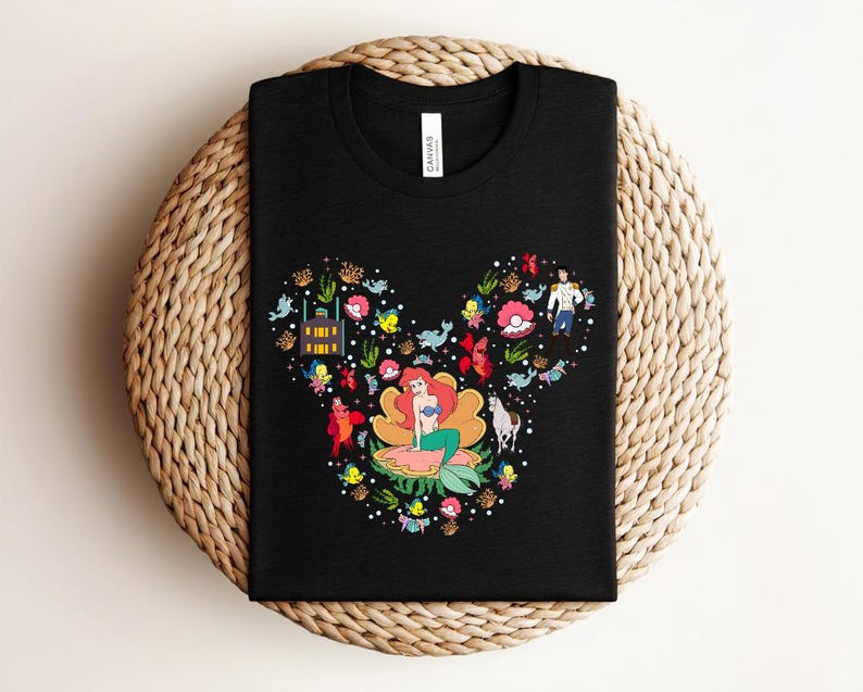 K&ouml;nnte beinhalten: Schwarzes T-Shirt mit einer farbenfrohen Grafik einer Meerjungfrau in einer Blume, umgeben von Meereskreaturen und Figuren. Das Design hat die Form eines M&auml;usekopfes. Das Shirt besteht aus einem weichen Material.