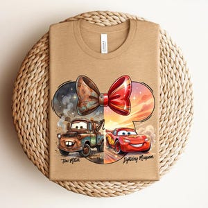 May include: A tan t-shirt featuring a graphic of a split bow design. The left side shows a rusty brown tow truck labelled Tow Mater, and the right side shows a red race car labelled Lightning McQueen. The design is set within a Mickey Mouse ear shape.