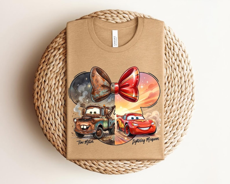May include: A tan t-shirt featuring a graphic of a split bow design. The left side shows a rusty brown tow truck labelled Tow Mater, and the right side shows a red race car labelled Lightning McQueen. The design is set within a Mickey Mouse ear shape.