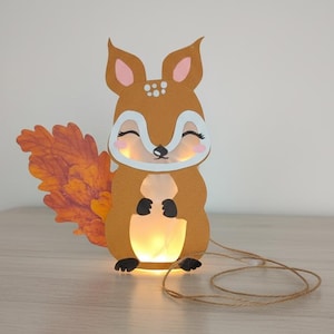 Może przedstawiać: Kapryśna, podświetlana latarnia w kształcie wiewiórki. Brązowa wiewiórka z papieru ma świecący środek, teksturowany pomarańczowy ogon i sznurek jutowy. Latarnia została zaprojektowana w kreskówkowym stylu, idealna do pokoi dziecięcych lub dekoracji sezonowych.