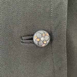 Puede incluir: Primer plano de un bot&oacute;n gris oscuro con un dise&ntilde;o floral y peque&ntilde;os acentos brillantes. El bot&oacute;n est&aacute; sujeto a una tela gris oscura, probablemente parte de un abrigo o chaqueta. El bot&oacute;n es redondo y tiene un patr&oacute;n detallado.