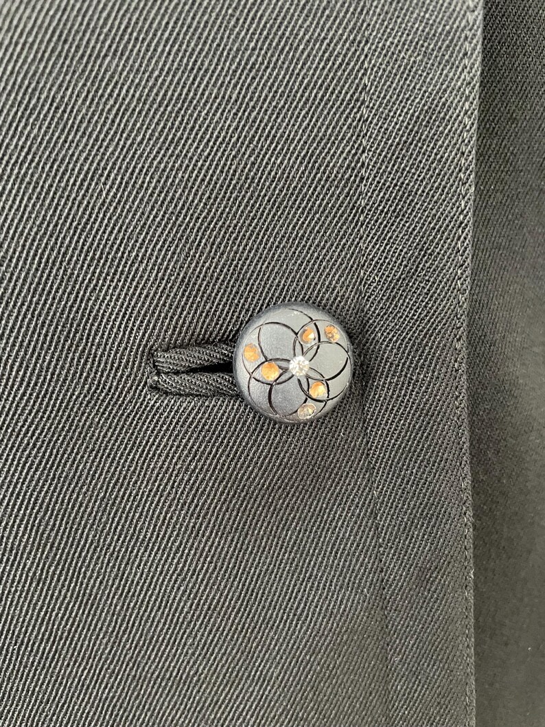 Puede incluir: Primer plano de un bot&oacute;n gris oscuro con un dise&ntilde;o floral y peque&ntilde;os acentos brillantes. El bot&oacute;n est&aacute; sujeto a una tela gris oscura, probablemente parte de un abrigo o chaqueta. El bot&oacute;n es redondo y tiene un patr&oacute;n detallado.