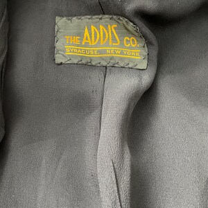 Puede incluir: Primer plano del interior de una prenda gris oscuro. Una etiqueta rectangular con el texto "THE ADDIS CO. SYRACUSE, NEW YORK" en letras amarillas est&aacute; cosida en el forro. La etiqueta tiene un borde verde.