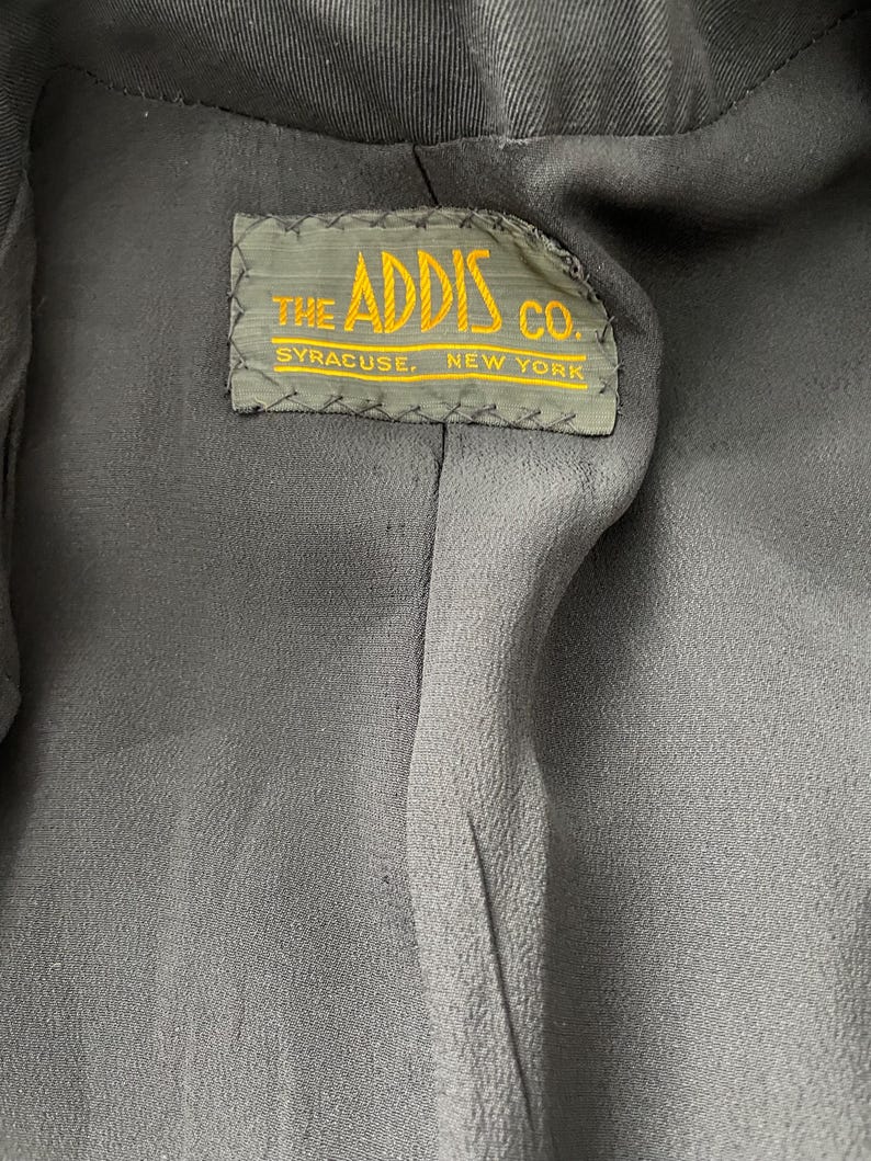 Puede incluir: Primer plano del interior de una prenda gris oscuro. Una etiqueta rectangular con el texto "THE ADDIS CO. SYRACUSE, NEW YORK" en letras amarillas est&aacute; cosida en el forro. La etiqueta tiene un borde verde.