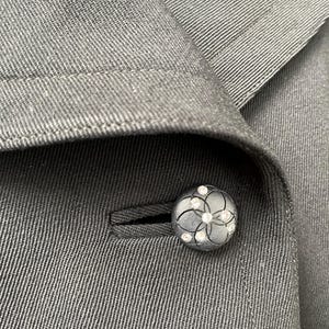 Puede incluir: Primer plano de una blazer gris oscuro con un bot&oacute;n decorativo. El bot&oacute;n es redondo, con un dise&ntilde;o floral y peque&ntilde;os adornos transparentes. La tela de la blazer tiene un patr&oacute;n sutil y texturizado.