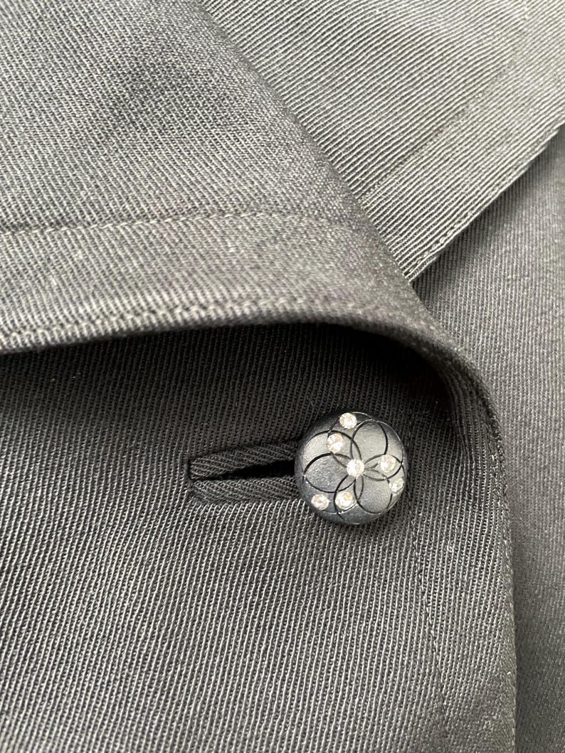 Puede incluir: Primer plano de una blazer gris oscuro con un bot&oacute;n decorativo. El bot&oacute;n es redondo, con un dise&ntilde;o floral y peque&ntilde;os adornos transparentes. La tela de la blazer tiene un patr&oacute;n sutil y texturizado.