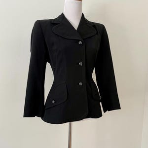 Puede incluir: Un blazer negro entallado, con cuello de solapa, mangas largas y botones decorativos. La chaqueta tiene dos bolsillos delanteros y se muestra en un maniqu&iacute;. El estilo es cl&aacute;sico y elegante.
