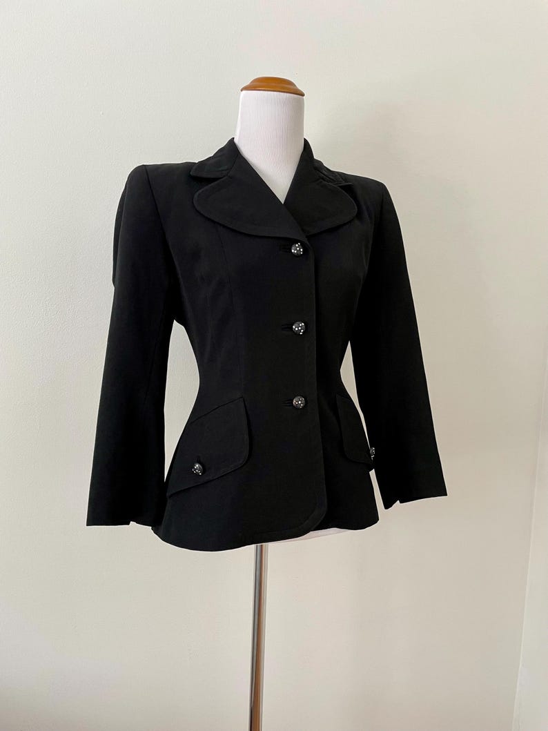 Puede incluir: Un blazer negro entallado, con cuello de solapa, mangas largas y botones decorativos. La chaqueta tiene dos bolsillos delanteros y se muestra en un maniqu&iacute;. El estilo es cl&aacute;sico y elegante.