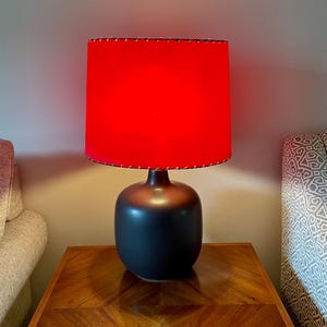 May include: A modern table lamp with a black base and a vibrant red lampshade. The lampshade is cylindrical and has a stitched trim. The lamp sits on a wooden table with a geometric pattern. The overall aesthetic is contemporary and stylish.