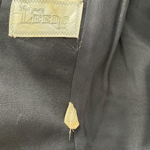 Puede incluir: Primer plano del interior de una prenda negra con una etiqueta que dice "that young LEEDS look". La etiqueta es de color crema con un borde cosido. Tambi&eacute;n es visible una etiqueta m&aacute;s peque&ntilde;a.