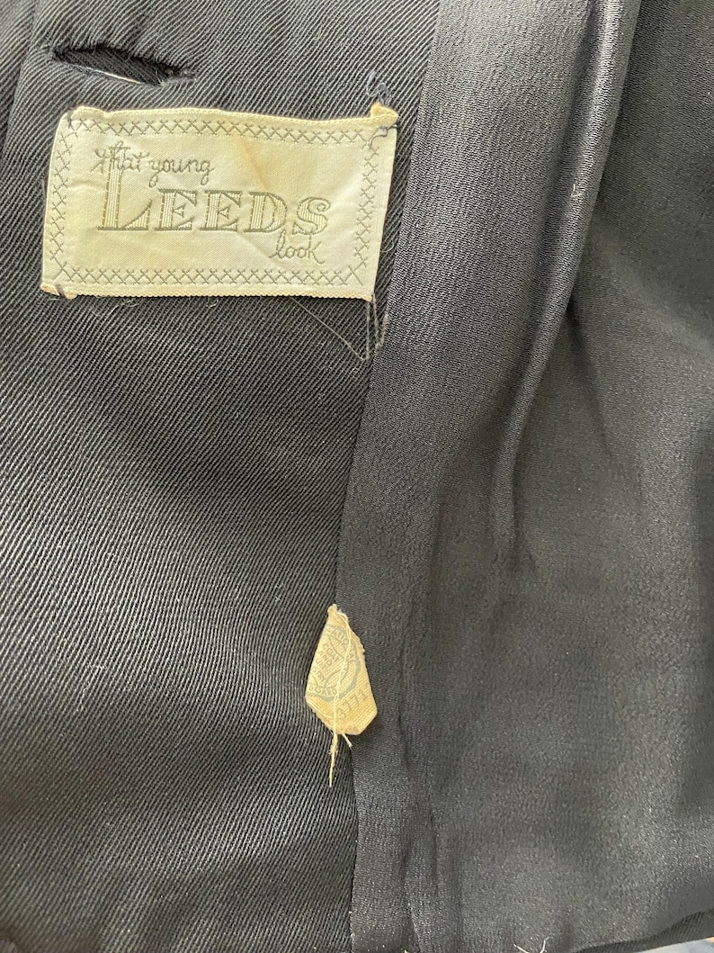 Puede incluir: Primer plano del interior de una prenda negra con una etiqueta que dice "that young LEEDS look". La etiqueta es de color crema con un borde cosido. Tambi&eacute;n es visible una etiqueta m&aacute;s peque&ntilde;a.