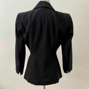 Puede incluir: Un blazer negro con un corte a medida, exhibido en un maniqu&iacute;. El blazer presenta solapas con muescas, mangas largas y una cintura entallada. La vista trasera destaca el dise&ntilde;o estructurado y la silueta cl&aacute;sica de la prenda.