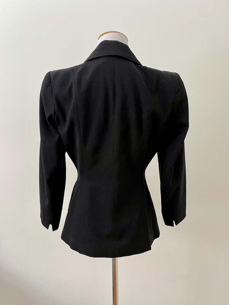 Puede incluir: Un blazer negro con un corte a medida, exhibido en un maniqu&iacute;. El blazer presenta solapas con muescas, mangas largas y una cintura entallada. La vista trasera destaca el dise&ntilde;o estructurado y la silueta cl&aacute;sica de la prenda.