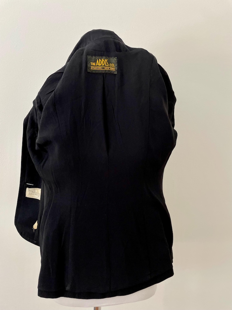 Puede incluir: Un blazer negro con un corte a medida. El interior presenta una etiqueta que dice "THE ADDIS CO. SYRACUSE, NEW YORK". El blazer tiene un dise&ntilde;o cl&aacute;sico, con solapas con muescas y cierre de dos botones. La tela parece ser un material liso y tejido.