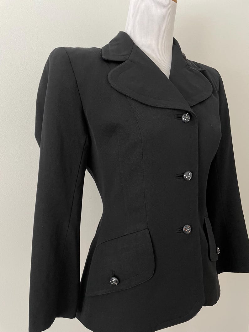 Puede incluir: Un blazer negro con cuello redondo, tres botones decorativos y dos bolsillos delanteros. El blazer se exhibe en un maniqu&iacute; y tiene un corte entallado. Los botones tienen un patr&oacute;n decorativo.