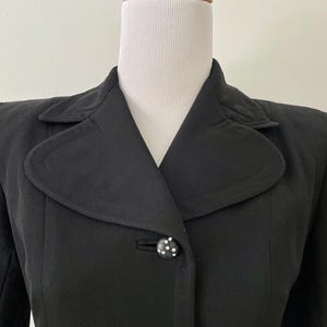 Puede incluir: Un blazer negro vintage con cuello con muescas y cierre de un solo bot&oacute;n. El bot&oacute;n es negro con lunares blancos. El blazer se exhibe en un maniqu&iacute; blanco.