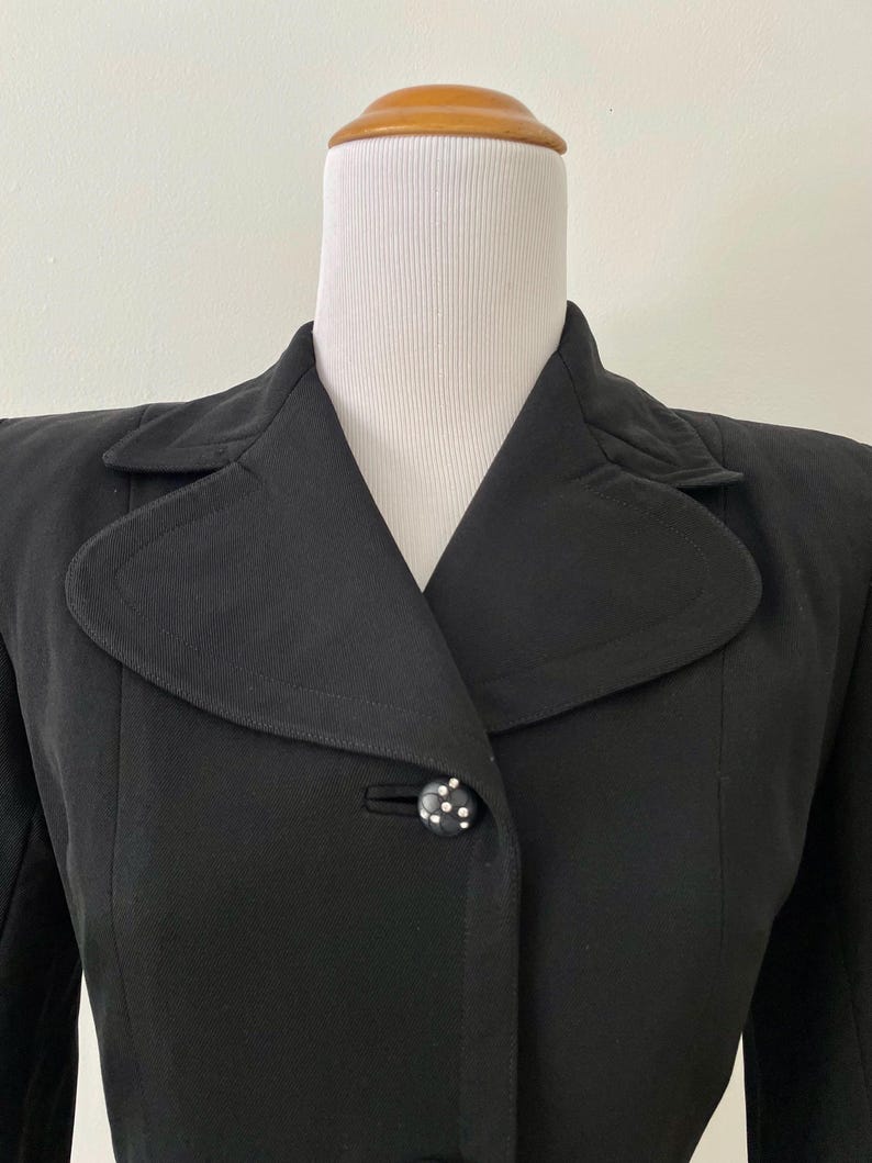 Puede incluir: Un blazer negro vintage con cuello con muescas y cierre de un solo bot&oacute;n. El bot&oacute;n es negro con lunares blancos. El blazer se exhibe en un maniqu&iacute; blanco.