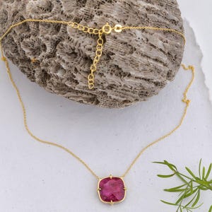 May include: A gold necklace with a square, magenta gemstone pendant. The pendant is set in a gold frame and suspended from a fine gold chain. The necklace is presented on a textured, light-coloured surface.