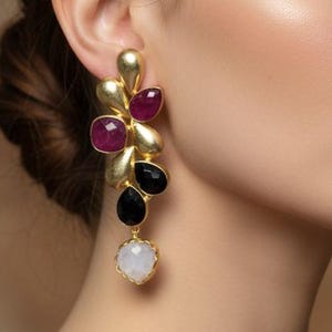 Puede incluir: Pendientes dorados llamativos con un diseño de racimo con acentos de piedras preciosas de rubí, negras y blancas. Los pendientes los lleva una persona con un vestido burdeos, creando un look sofisticado. Un accesorio audaz.