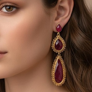 Puede incluir: Una mujer lleva pendientes colgantes dorados con grandes piedras preciosas rojas en forma de lágrima. Los pendientes tienen un diseño detallado de filigrana alrededor de las piedras preciosas. La mujer tiene el pelo largo y ondulado de color marrón.
