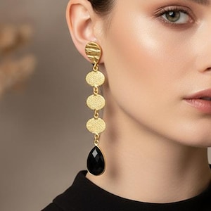 May include: Gold-tone drop earrings with a textured disc design and a black teardrop-shaped stone. The earrings are long and dangle, adding a touch of elegance. The model is wearing a black top.