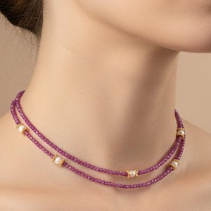 May include: A two-strand necklace with small, deep pink beads and gold-coloured accents. The necklace is draped around the neck, highlighting its delicate design and colour contrast. The jewellery is a fashion accessory.