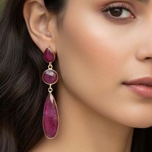 Könnte beinhalten: Elegante goldfarbene Ohrringe mit drei rubinroten Edelsteinen. Der obere Stein ist tropfenförmig, gefolgt von einem runden Stein und einem größeren Tropfen unten. Die Ohrringe sind ein auffälliges Accessoire.