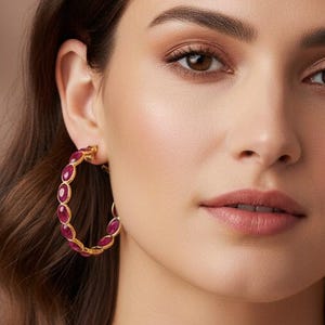 May include: Gold-tone hoop earrings with a row of oval-shaped, deep red gemstones. The earrings are worn by a person with brown hair and a neutral-toned top. The background is a soft, warm color.