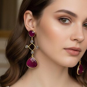 May include: Gold-coloured earrings with large, faceted, deep red gemstones. The earrings feature a geometric design with smaller grey stones and a teardrop-shaped ruby. The woman in the image is wearing a cream-coloured dress.