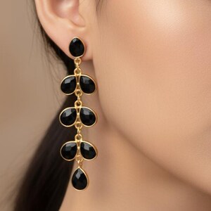 Peut inclure: Boucles d'oreilles dorées élégantes avec des gouttes de pierres précieuses noires. Les boucles d'oreilles présentent une série de pierres noires en forme de goutte suspendues à une chaîne dorée. Portées par une personne aux cheveux foncés.