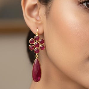 May include: Gold-tone earrings featuring a floral cluster design with ruby-red gemstones. Each earring has a teardrop-shaped ruby pendant. The earrings are worn by a person with dark hair and a neutral background.