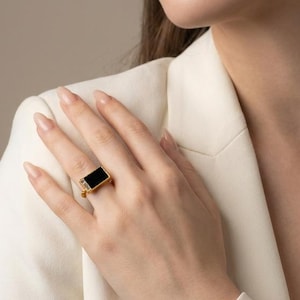 May include: A gold ring featuring a rectangular black stone set in a polished metal band. The ring is worn on a finger, showcasing the contrast between the dark stone and the metallic gold setting, which has a clean, geometric design.