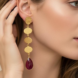 Könnte beinhalten: Eine Frau trägt einen langen goldenen Ohrring mit einem tropfenförmigen, roten Edelstein. Der Ohrring hat eine Reihe von vier goldenen ovalen Formen, wobei die oberste Form ein strukturiertes Design aufweist. Die Frau berührt ihr Ohr, und der Hintergrund ist verschwommen.