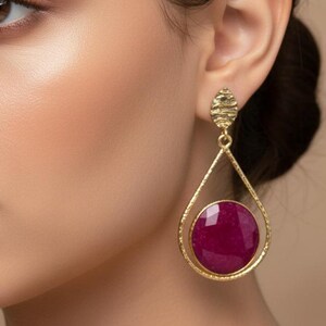 Può includere: Orecchini a goccia color oro con un design strutturato. Ogni orecchino presenta una grande gemma rotonda di colore rosso intenso incastonata in una cornice delicata. Gli orecchini sono esposti su un orecchio, mostrando il loro design elegante.