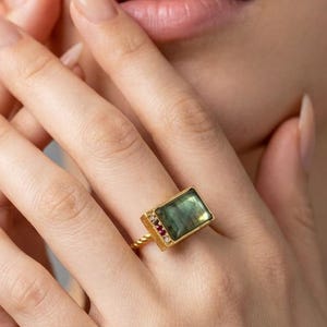 May include: A gold ring featuring a large rectangular green gemstone set in a bezel. The band has a twisted texture, and the side of the setting is accented with three small, colorful stones, including a red stone and two clear stones.