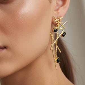 Könnte beinhalten: Goldfarbene Ohrringe mit einem linearen, geometrischen Design. Die Ohrringe weisen eine Reihe sich kreuzender Linien und drei schwarze, kreisförmige Akzente auf. Die Ohrringe sind hängend und verleihen der Schmuckkollektion eine moderne Note.