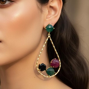 May include: Gold-toned teardrop earrings with a cluster of colorful gemstones. The earrings feature a square green stone at the top, with a teardrop-shaped frame holding a black, red, and green stone cluster. The earrings are a statement piece.