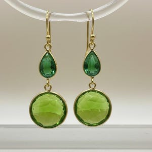 May include: A pair of gold-coloured drop earrings. Each earring features a teardrop-shaped green gemstone above a large, round, faceted green gemstone. The earrings are suspended from a clear, curved display stand.