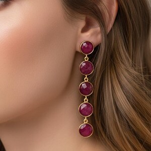 May include: A close-up of a person wearing long, dangling earrings. The earrings feature five round, deep red gemstones set in gold-coloured metal. The gemstones are evenly spaced and connected by delicate chains.