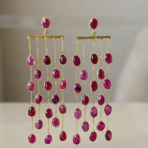 以下が含まれることがあります： 繊細なゴールドチェーンから吊り下げられた、楕円形の濃いピンク色の宝石が連なったドロップイヤリング。 イヤリングは、上部に小さな丸い宝石と長方形のゴールドバーが特徴です。 中立的な背景に設定されています。