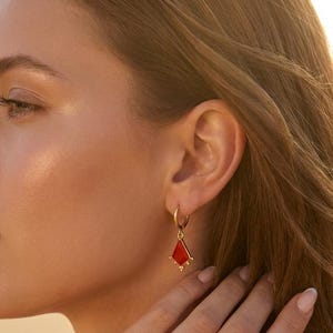 May include: A gold hoop earring featuring a dangling kite-shaped charm with a vibrant red enamel inlay. The charm is accented with small gold beads at the bottom point and along the sides, suspended from a thin, circular gold hoop wire.