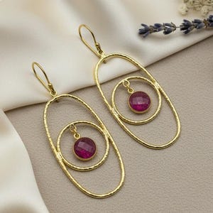 May include: A pair of gold-colored earrings with a unique design. Each earring features an oval frame with a smaller, concentric oval inside, and a round, magenta-colored gemstone suspended in the center. The earrings are displayed on a white satin surface.
