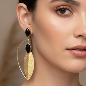 Puede incluir: Una mujer lleva pendientes dorados con detalles negros. Los pendientes tienen forma de lágrima con superficie texturizada y una piedra negra en la parte superior e inferior. Joyería.